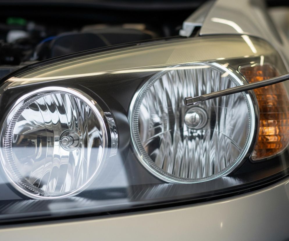 Przednia lampa samochodowa, dominująca na zdjęciu, posiada trzy okrągłe elementy świetlne: dwa główne reflektory z przezroczystymi kloszami i srebrnymi obwódkami oraz mniejszy, pomarańczowy kierunkowskaz po prawej stronie. Na tle prawej głównej lampy widoczny jest koniec srebrnego, metalowego narzędzia, które delikatnie dotyka jej powierzchni. Nad lampą, z lewej strony, uchylona jest maska samochodu, ukazując fragmenty silnika.