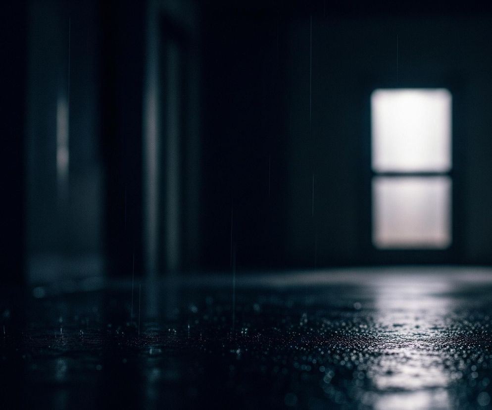 Na pierwszym planie widoczne jest ciemne, błyszczące podłoże pokryte licznymi kroplami wody, z wyraźnym rozmyciem perspektywy w kierunku horyzontu. W lewym dolnym rogu majaczą rozmyte, czerwone i niebieskie światła, podczas gdy centralnie na dalszym planie znajduje się jasne, prostokątne okno lub otwór, którego światło odbija się od mokrej powierzchni. Całość jest utrzymana w ciemnej tonacji z dominującymi odcieniami granatu i czerni, tworząc wrażenie deszczu padającego wewnątrz lub na otwartej przestrzeni oświetlonej jedynie odległym źródłem światła.