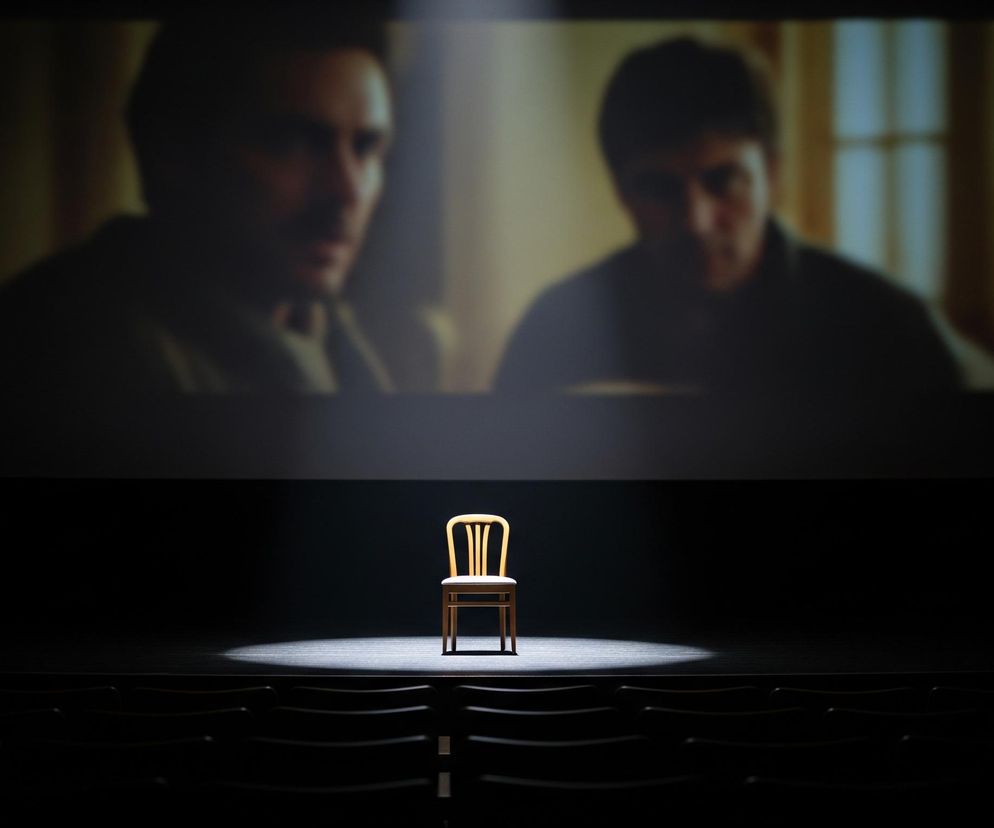 Na środku ciemnej sceny, pod pojedynczym snopem światła, stoi drewniane krzesło z jasnym siedziskiem. Za krzesłem, na dużej, rozmazanej projekcji filmowej widoczne są niewyraźne sylwetki dwóch mężczyzn w stonowanych kolorach, z tłem w odcieniach beżu i błękitu. Poniżej sceny, w głębokim cieniu, zarysowane są rzędy pustych siedzeń kinowych, co sugeruje ciemne wnętrze sali.