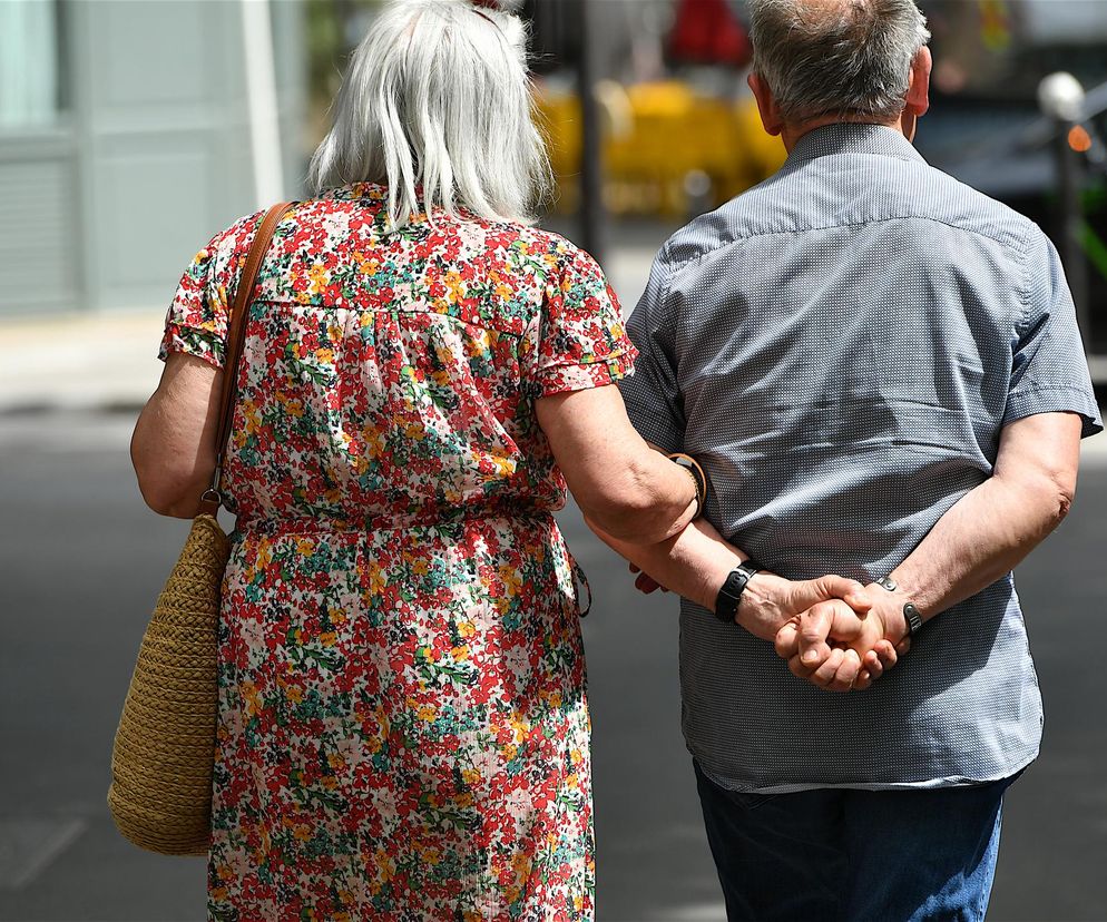 Para seniorów idąca ulicą, kobieta w kwiecistej sukience i z torebką na ramieniu, mężczyzna w jasnej koszuli z krótkim rękawem. Zdjęcie symbolizuje wsparcie i wspólne pokonywanie trudności, nawiązując do dopłat dla seniorów, o których przeczytasz na Poradnik Zdrowie.