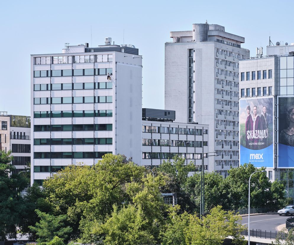 Modernistyczny budynek Solec ZEITRAUM Student Housing w Warszawie, z charakterystyczną białą elewacją i pasmami okien, odzwierciedla projekt pracowni Grupa 5 Architekci. W górnej części widoczny pracownik konserwacji, co jest częścią cyklu życia w architekturze, o czym można przeczytać na Architektura Murator Plus.