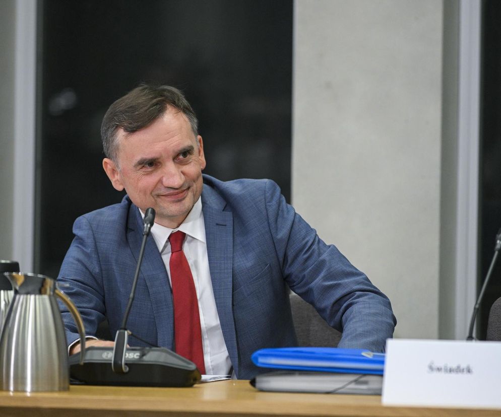 Zbigniew Ziobro w niebieskiej marynarce i czerwonym krawacie, siedzący przy stole podczas posiedzenia komisji śledczej do spraw Pegasusa, uśmiecha się. W tle widać fragmenty sali, a na stole termosy. Więcej informacji o zabezpieczeniu majątku Ziobry na Super Biznes.