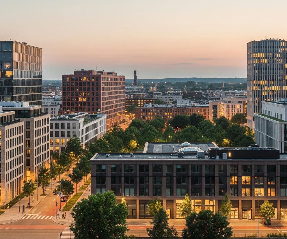 Panorama miejska oświetlona wieczornym, złocisto-różowym światłem, prezentująca architekturę miejską w otoczeniu zieleni. Dominują wysokie, nowoczesne budynki o różnej kolorystyce i fakturze, z których wiele ma podświetlone wnętrza, widoczne przez liczne okna. W dolnej części obrazu widoczna jest ulica z przejściami dla pieszych, drzewami wzdłuż chodnika oraz kilkoma przechodniami. W oddali rozpościera się krajobraz miejski z innymi budowlami i wieżą radiową na horyzoncie, a nad całością rozciąga się bezchmurne niebo.