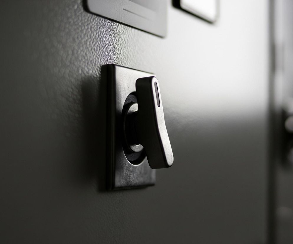 Centralnym elementem jest metaliczny przełącznik z zaokrągloną dźwignią, osadzony w prostokątnej ramie na ciemnoszarym, lekko chropowatym panelu. Dźwignia skierowana jest lekko w prawo i do góry, a jej górna krawędź jest jaśniejsza od reszty, co wskazuje na padające światło. W tle, częściowo widoczne są inne elementy panelu, również w odcieniach szarości, ale są one rozmyte i nieostre. Całość utrzymana jest w stonowanej kolorystyce szarości i czerni, z wyraźnym światłocieniem, dodającym głębi.