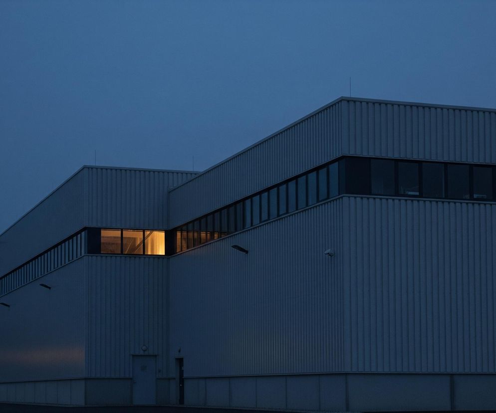 Na obrazie dominuje duży, prostokątny budynek przemysłowy o szarych, pionowo żebrowanych ścianach, widziany z perspektywy nocnej lub o zmierzchu. Niebo jest granatowe, przechodzące w ciemny błękit. W górnej części budynku znajduje się pas ciemnych okien, z których jedno, umieszczone w centrum, świeci intensywnym, ciepłym, żółto-pomarańczowym światłem. Poniżej okien widać ciemne drzwi i niewielki daszek nad nimi, a także inne detale architektoniczne i elementy oświetlenia zewnętrznego.
