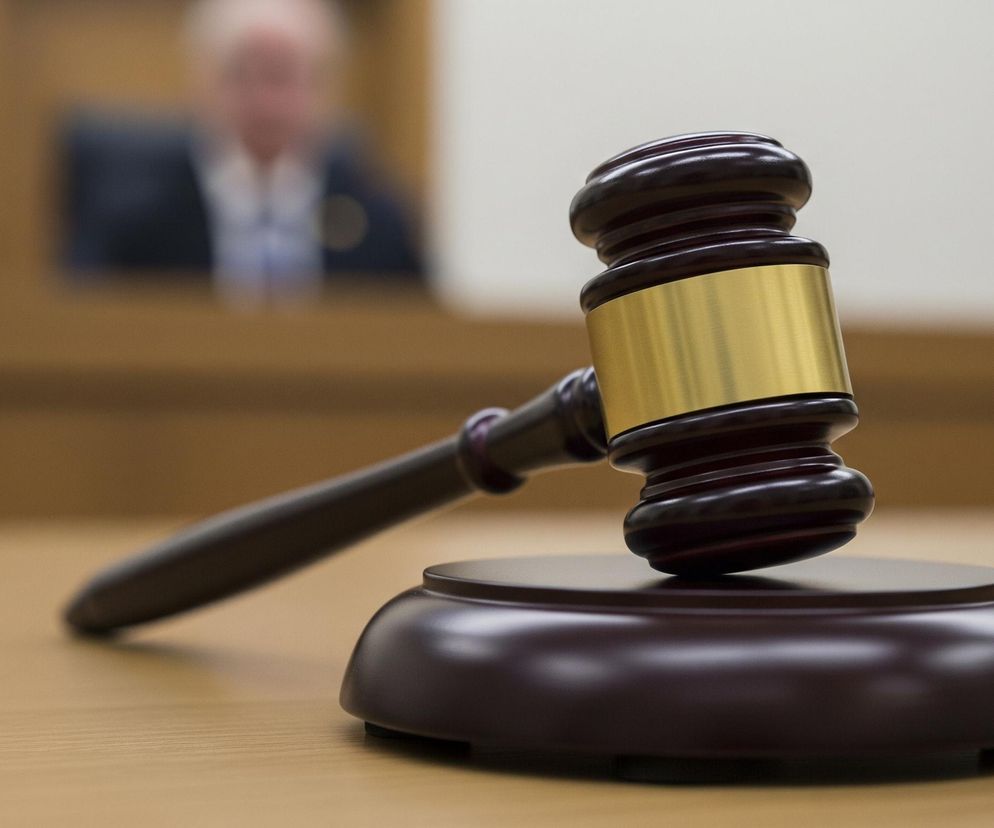 Młotek sędziowski wykonany z ciemnego drewna, ze złotym pierścieniem na główce, leży na okrągłej podstawce z tego samego materiału. Całość spoczywa na jasnobrązowym, drewnianym stole. W tle, rozmyta postać osoby w garniturze i koszuli widoczna jest za drewnianą balustradą. Scena rozgrywa się w pomieszczeniu z jasną ścianą w tle.