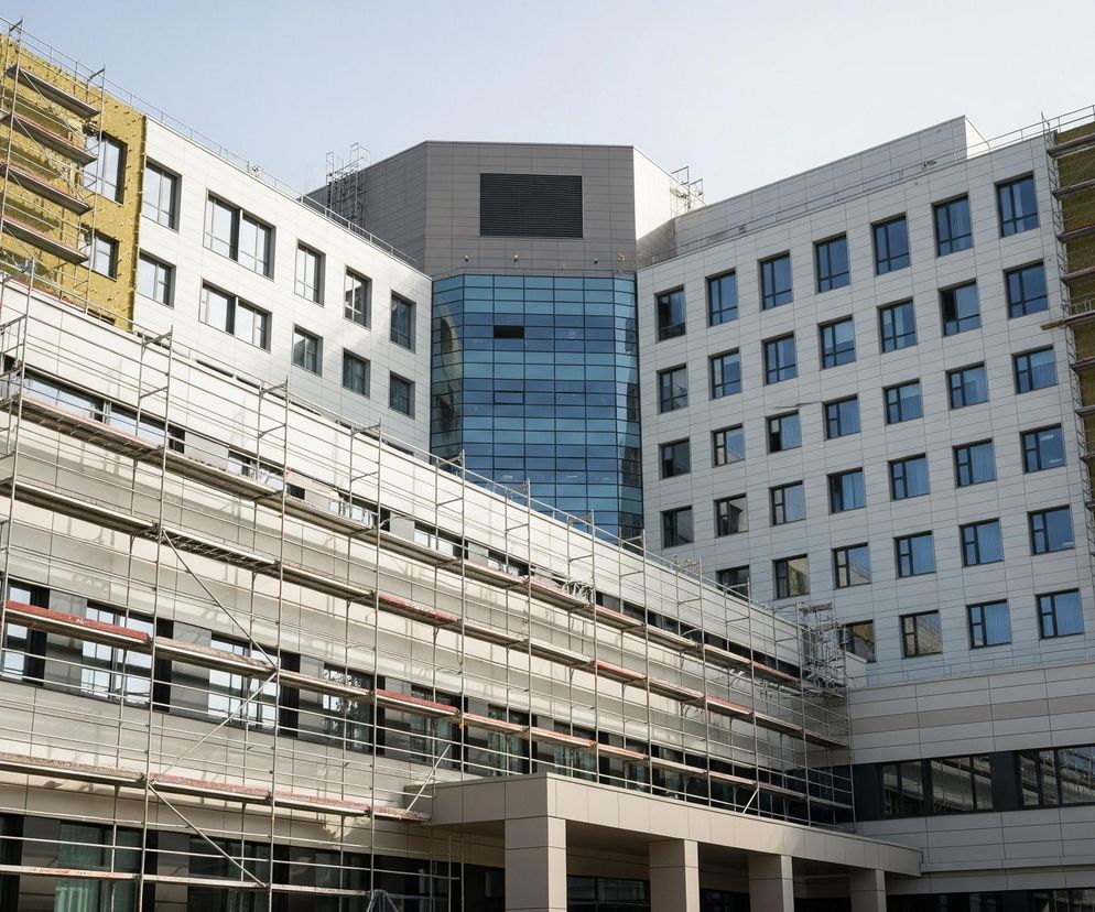 Duży, wielopiętrowy budynek w trakcie budowy lub remontu, z widocznymi rusztowaniami wokół części elewacji. Centralna bryła budynku wyróżnia się przeszkloną fasadą w odcieniach błękitu, otoczoną dwoma skrzydłami w jasnych, beżowych panelach. Na bocznych ścianach skrzydeł, jak i na części frontowej, widoczne są warstwy żółtej izolacji termicznej, pokrytej konstrukcją rusztowania. Niebo nad budynkiem jest jasne i bezchmurne.