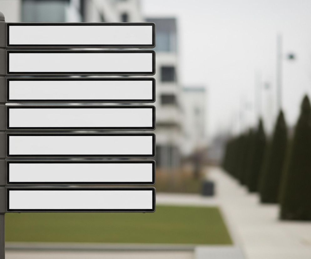 Szara tablica informacyjna z siedmioma pustymi, białymi prostokątami na ciemnym tle, umieszczona jest po lewej stronie kadru. Tablica ma zaokrąglone górne zakończenie i przymocowana jest do pionowego słupa. W tle widoczne są rozmyte, nowoczesne budynki o jasnych elewacjach, a także pas zielonej trawy na pierwszym planie po lewej. Po prawej stronie, w oddali, rozciąga się chodnik z rzędem stożkowatych, ciemnozielonych krzewów i pojedynczymi, nagimi drzewami oraz słupami oświetleniowymi, wszystko w stonowanych barwach.
