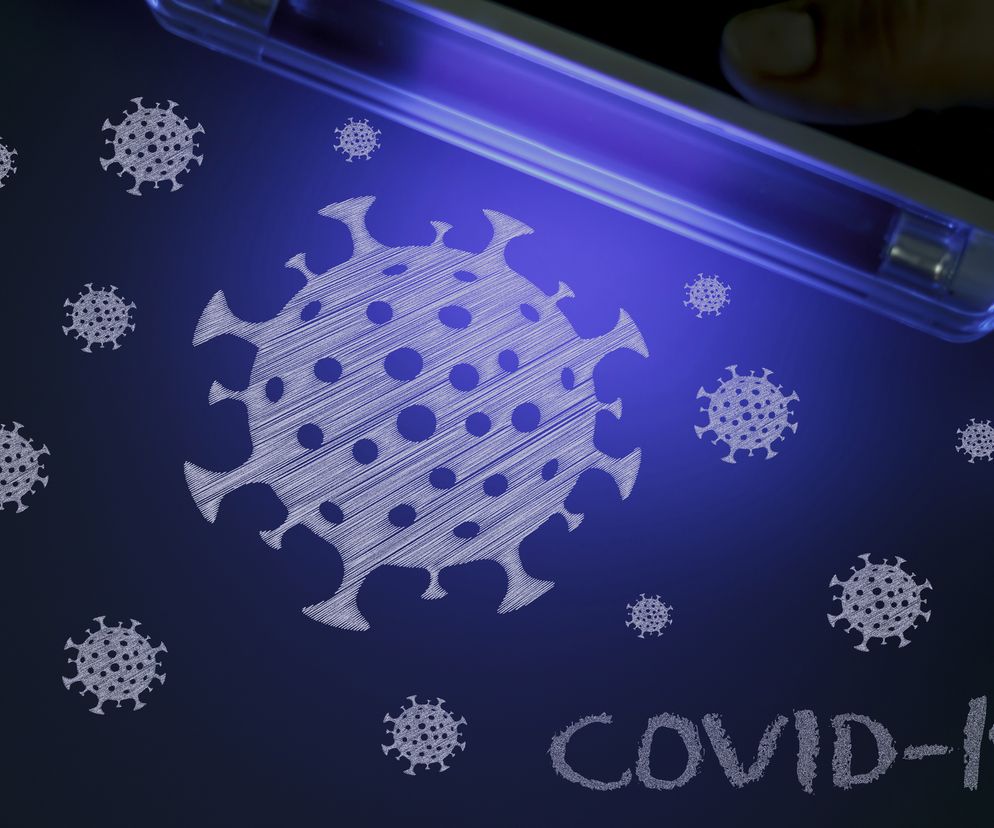 Grafika przedstawiająca dłoń trzymającą lampę UV, oświetlającą rysunki wirusów koronawirusa i napis COVID-19 na ciemnym tle. Ilustracja symbolizuje badanie i walkę z pandemią, a więcej na ten temat przeczytasz na Poradnik Zdrowie.