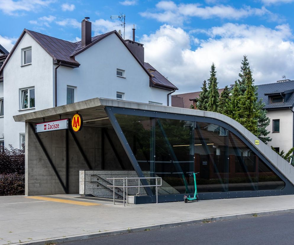Nowoczesne wejście do stacji metra Zacisze w Warszawie z logo metra i nazwą stacji, betonowa konstrukcja i szklane ściany kontrastują z tradycyjnymi domami jednorodzinnymi i zielonymi drzewami w tle. Więcej o nietypowych osiedlach na Architektura Murator Plus.