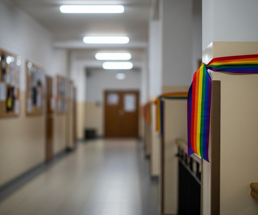 W centrum kadru znajduje się długi korytarz, oświetlony rzędami poziomych, prostokątnych lamp sufitowych. Po prawej stronie, na pierwszym planie, widoczna jest tęczowa wstążka, zwisająca z beżowego słupa lub ściany. Dalej wzdłuż korytarza, na kolejnych słupach, zauważalne są podobne tęczowe wstążki, tworzące powtarzający się wzór. Po lewej stronie korytarza widoczne są tablice korkowe z przypiętymi ogłoszeniami i kartkami. Na końcu korytarza znajduje się dwuskrzydłowe, drewniane drzwi.