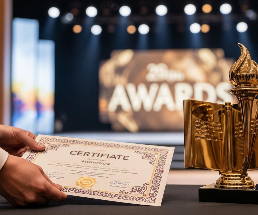 Mężczyzna w eleganckim, ciemnym garniturze z białą koszulą i mankietami, trzyma w dłoniach biały certyfikat z ozdobnymi, brązowymi ramkami i napisem CERTIFICATE. Obok, na ciemnym blacie, stoi złota statuetka, stylizowana na otwartą księgę z płomieniem na szczycie, umieszczona na czarnej, prostokątnej podstawie. Tło stanowi rozmyta scena, prawdopodobnie duża sala lub scena wydarzenia, z widocznym, rozmytym napisem AWARDS oraz jasnymi punktami świetlnymi.