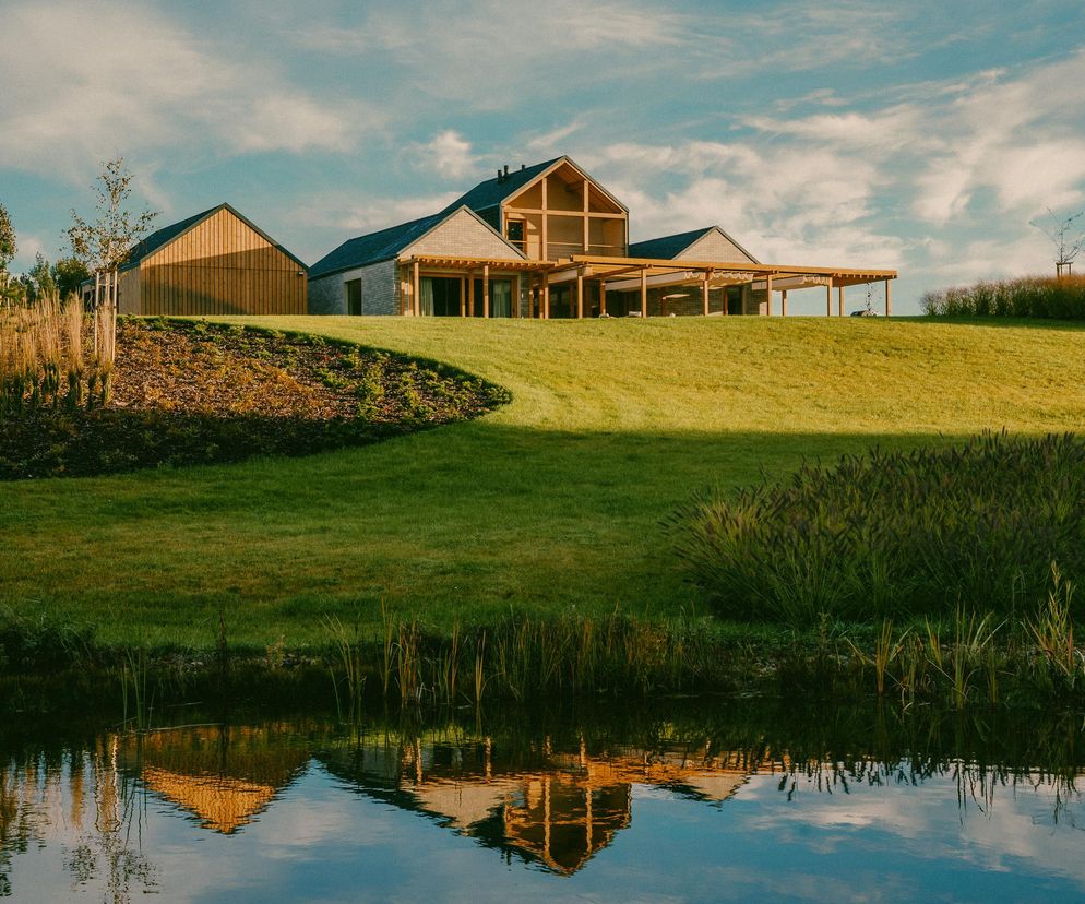 Zespół domów letniskowych Pasym Osada projektu Noke Architects, widoczny na zielonym wzgórzu, z odbiciem w wodzie na pierwszym planie. Architektura Murator Plus prezentuje zgłoszenie do konkursu Życie w Architekturze 2025, wyróżniające się połączeniem tradycji Mazur z nowoczesnymi rozwiązaniami prefabrykowanymi.