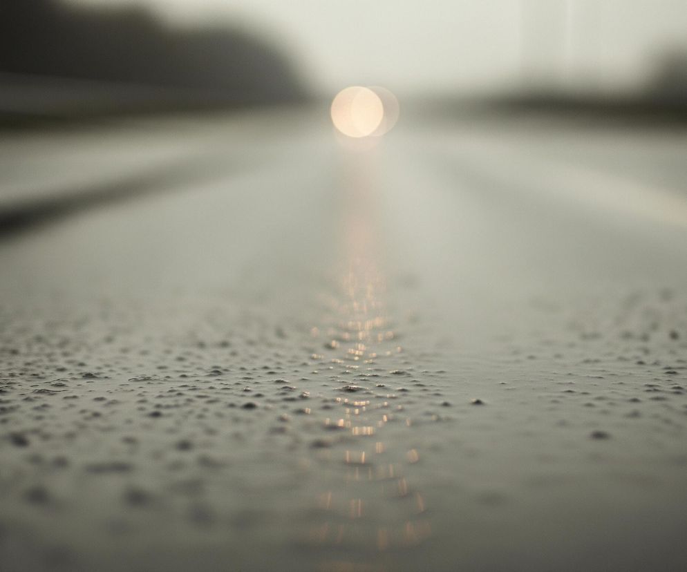Mokra, szara nawierzchnia drogi zajmuje większą część kadru, z wyraźnymi kroplami wody na pierwszym planie. W oddali widać rozmyte, jasne światło, które odbija się jako pionowa smuga na mokrej powierzchni. Po prawej stronie widoczna jest rozmyta, jasna linia pozioma, prawdopodobnie pas ruchu.