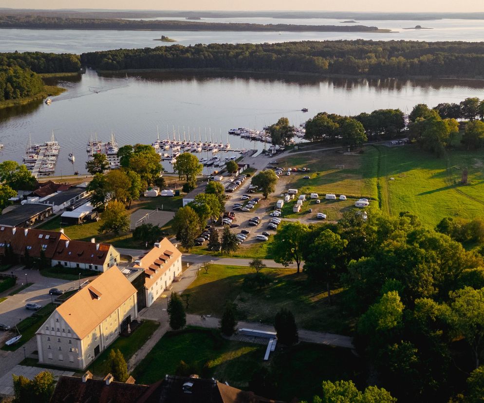 Multimedialne Centrum Żeglarstwa i Ekologii Memuak w Sztynorcie, z lotu ptaka. Widoczne zabytkowe budynki z rudymi dachami, marina z licznymi jachtami, camping oraz otaczająca jezioro bujna zieleń. O projekcie centrum przeczytasz na Architektura Murator Plus.