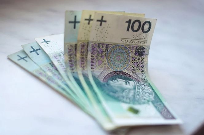 Stos banknotów o nominale 100 złotych, ułożonych w wachlarz, symbolizuje kwestie finansowe. Obraz nawiązuje do tematu obniżek stóp procentowych i ich wpływu na kredyty, co jest kluczowe dla czytelników Super Biznes.