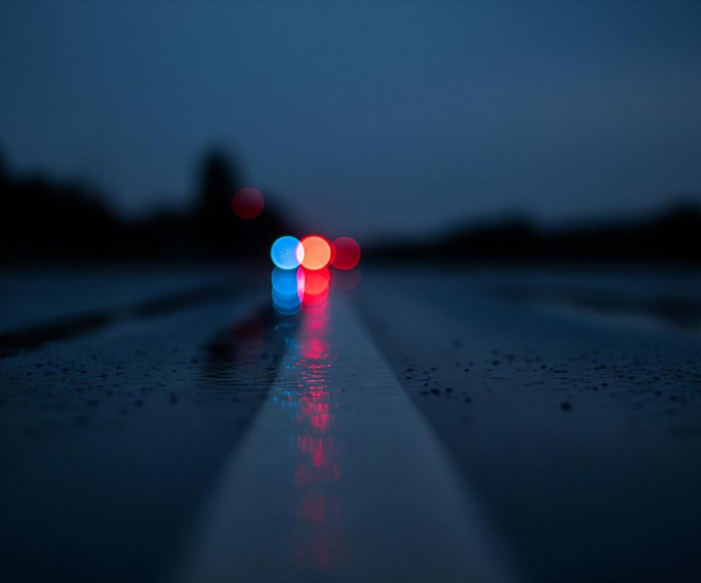 Mokra, ciemna droga z widocznym białym pasem pośrodku rozciąga się w głąb obrazu. Na horyzoncie, zza ciemnego zarysu drzew lub krzewów, migoczą rozmyte światła w kolorach czerwonym i niebieskim. Światła te, a także biały pas, odbijają się wyraźnie w kałużach na powierzchni drogi, tworząc symetryczne refleksy. Całość utrzymana jest w ciemnych, nocnych barwach, z dominującym granatem i odcieniami szarości.