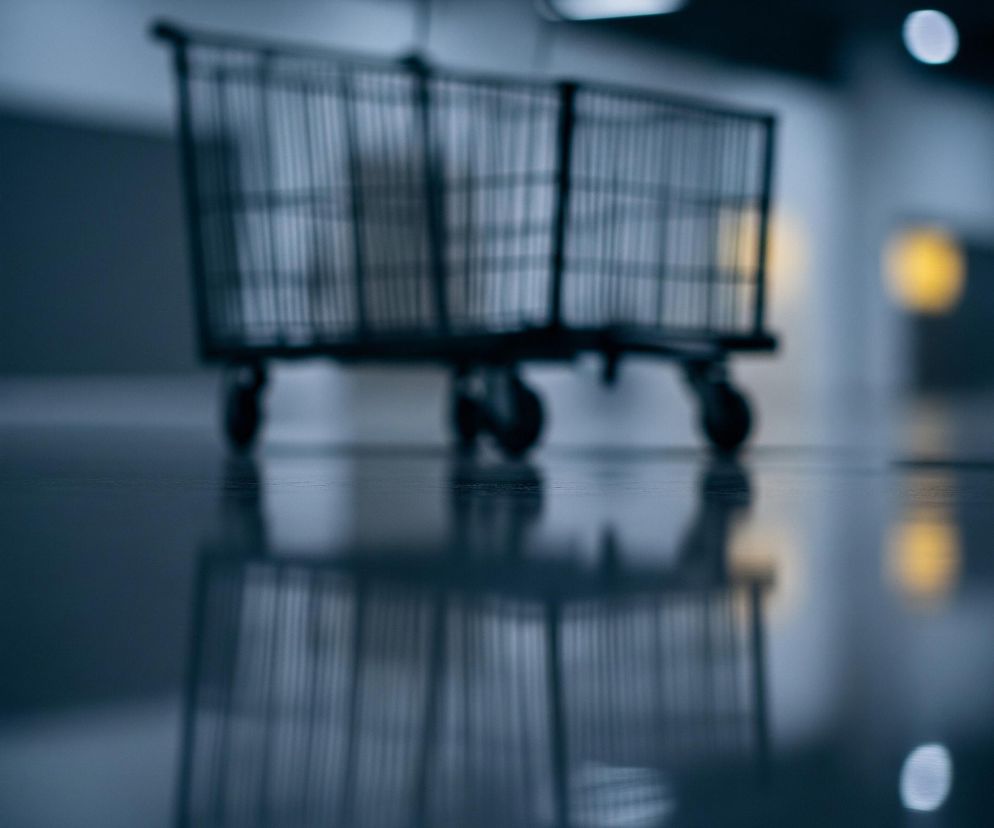 Na pierwszym planie, centralnie, widać metalowy wózek na zakupy, lekko rozmyty, z wyraźnymi pionowymi prętami tworzącymi jego kosz. Wózek odbija się w ciemnej, błyszczącej podłodze, co tworzy lustrzany efekt. Tło jest nieostre, dominują na nim odcienie szarości i błękitu, z kilkoma jasnymi, okrągłymi punktami światła, oraz jednym wyraźnym, żółtym punktem światła po prawej stronie. Ogólna atmosfera jest stonowana i nieco melancholijna.