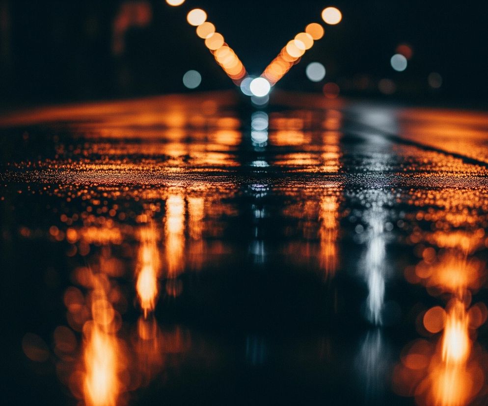 Droga w nocy, pokryta kałużami, odbija rozmyte światła uliczne. Jasnopomarańczowe i białe punkty świetlne tworzą efekt bokeh na górze kadru oraz ich wydłużone odbicia na mokrej nawierzchni. Czarne tło dominujące w górnej części obrazu pogłębia wrażenie ciemności, podkreślając świetliste refleksy.
