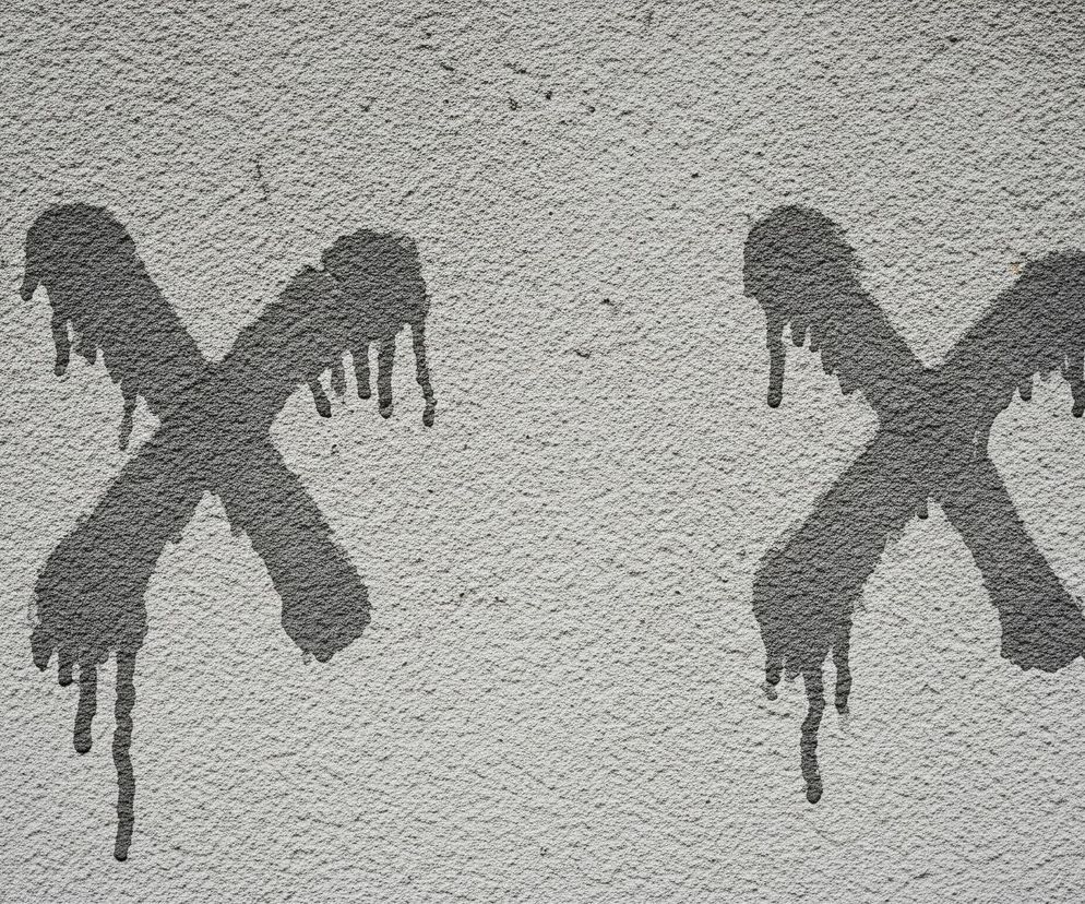 Na szarej, teksturowanej ścianie, pokrytej nierówną, ziarnistą fakturą, widoczne są dwa stylizowane, ciemnoszare znaki X. Oba znaki wyglądają jak wykonane za pomocą farby lub sprayu, z wyraźnymi zaciekami i kroplami spływającymi w dół z ich ramion, co sugeruje świeżość lub płynność substancji. Znaki są rozmieszczone obok siebie, centralnie na tle ściany, z lekkimi śladami brudu i niewielkimi, ciemniejszymi plamkami na jej powierzchni.