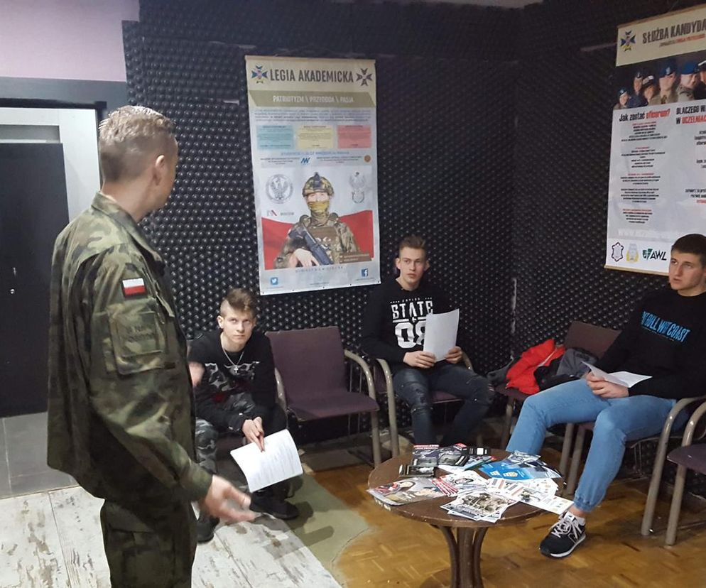 Wojskowy w mundurze z polską flagą na ramieniu rozmawia z trzema młodymi mężczyznami, którzy siedzą na krzesłach, trzymając dokumenty. Na ścianie wiszą plakaty Legia Akademicka oraz Służba Kandydacka. Więcej na temat obowiązków wojskowych przeczytasz na Super Biznes.