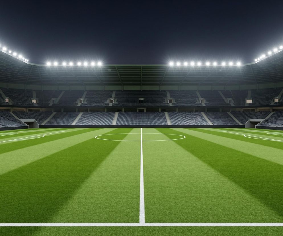 Oświetlony stadion sportowy z zielonym boiskiem zajmuje większą część obrazu. Boisko, podzielone białymi liniami, ma jaśniejsze i ciemniejsze pasy trawy, biegnące prostopadle do linii bocznych. W centrum boiska znajduje się białe koło, a po obu stronach boiska widoczne są bramki składające się z siatki oraz podwójnych, wysokich słupów z czerwonymi końcówkami. Trybuny stadionu, wypełnione rzędami pustych, ciemnych siedzeń, otaczają boisko, a nad nimi, pod zadaszeniem, świecą rzędy jasnych lamp, intensywnie oświetlających całą przestrzeń. Ciemne, jednolite niebo widoczne jest ponad konstrukcją stadionu.