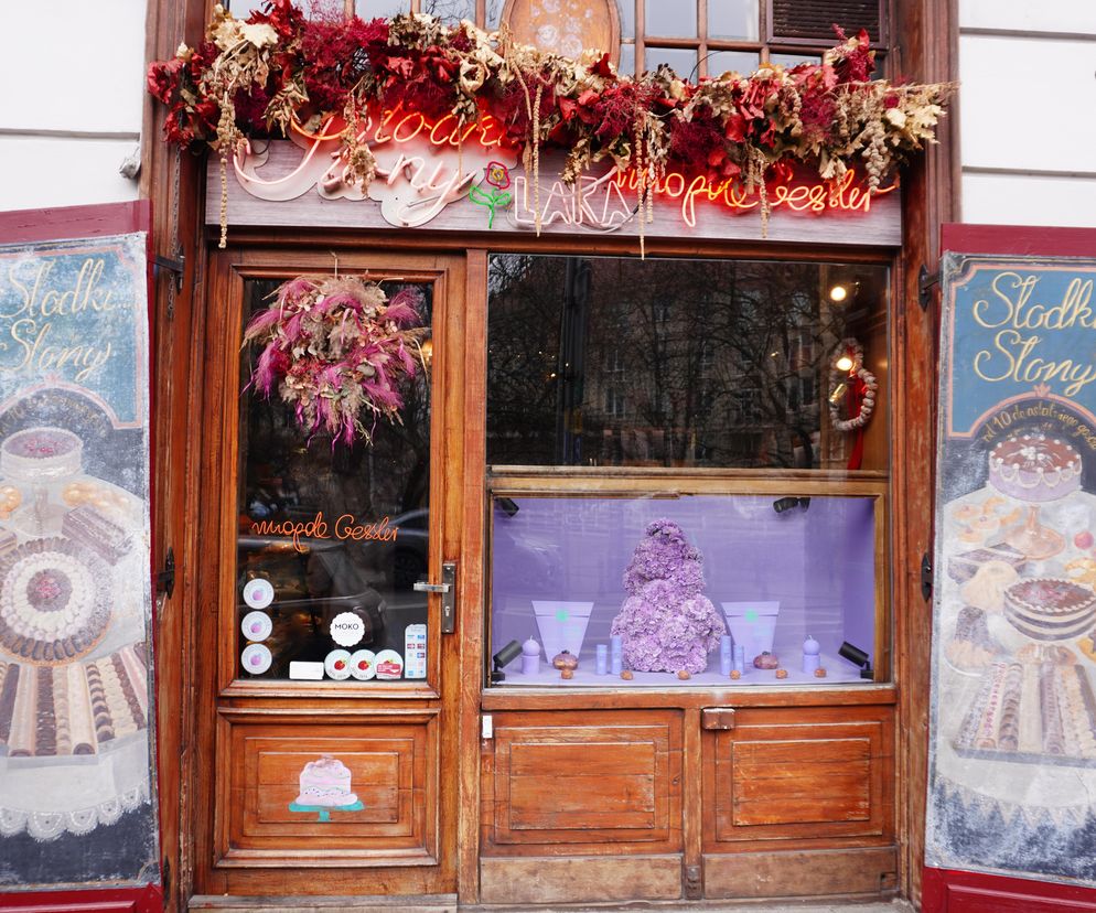 Wejście do cukierni Słodki Słony Magdy Gessler z drewnianymi drzwiami i witryną, ozdobioną neonowym napisem i suszonymi kwiatami. Obok malowane tablice z motywami słodkości. O aktualnych cenach pączków dowiesz się z artykułu na Super Biznes.