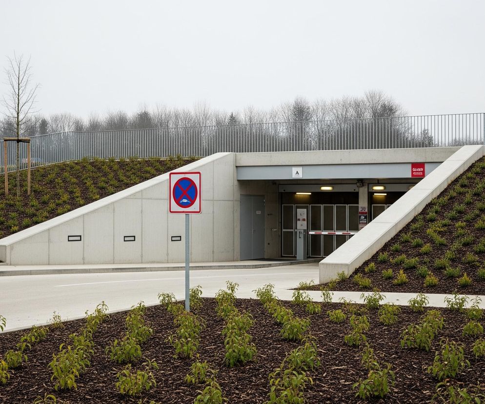 Wjazd do parkingu podziemnego o szarych betonowych ścianach znajduje się centralnie, otoczony zielonymi skarpami porośniętymi młodymi roślinami na ciemnej ściółce. Przed wjazdem, na środku szarego chodnika, stoi znak zakazu zatrzymywania się i postoju. Nad wjazdem widoczne są litera A oraz czerwony szyld z białym napisem. Poziomy szlaban częściowo blokuje wjazd, a w tle widać rzędy nagich drzew oraz fragmenty szarego ogrodzenia.