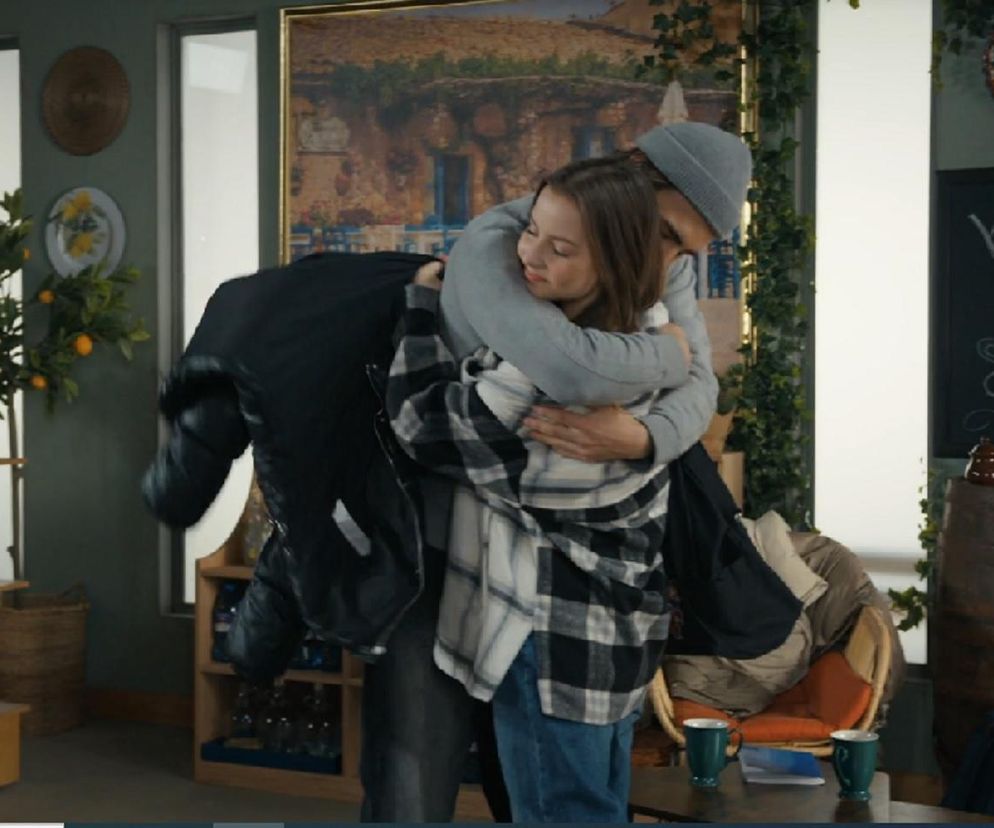 Młoda kobieta, Ewa (Gabriela Ziembicka), w objęciach mężczyzny, Romeo (Jacek Tyszkiewicz), oboje uśmiechnięci. Scena z serialu Barwy szczęścia, gdzie ich relacja jest centralnym punktem fabuły, o czym można przeczytać na Super Seriale. W tle wnętrze z półkami na wino i elementami dekoracyjnymi.