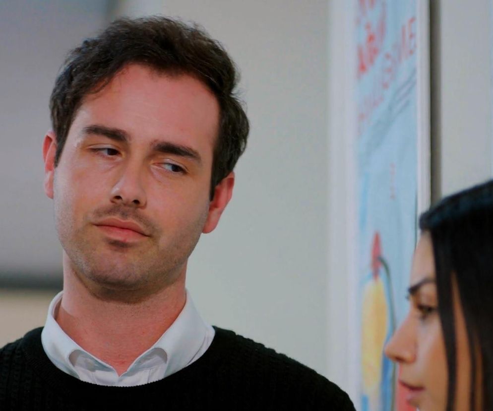 Levent, mężczyzna o ciemnych włosach i zamyślonym wyrazie twarzy, w białej koszuli i czarnym swetrze, obok niego fragment postaci Bahar. Scena z serialu Miłość i nadzieja, gdzie Levent i Bahar planują zniszczenie telefonu. Więcej o ich losach na Super Seriale.