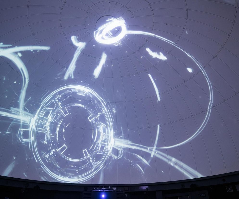 Kopuła planetarium jest głównym obiektem zdjęcia, wypełniając jego górną i środkową część. Na jej powierzchni, podzielonej na panele przez ciemniejsze linie, wyświetlana jest projekcja złożona z jasnych, świecących linii i kształtów w odcieniach bieli i jasnego błękitu, przypominających abstrakcyjne wzory technologiczne lub energetyczne. W centralnej części projekcji dominuje duży okrągły wzór z mniejszymi, prostokątnymi elementami wewnątrz, a wokół niego rozchodzą się falujące, dynamiczne smugi. Dolna część kadru to ciemna, pozioma powierzchnia widowni lub bazy projekcyjnej, na której widać niebieskie światło wychodzące z projektora.
