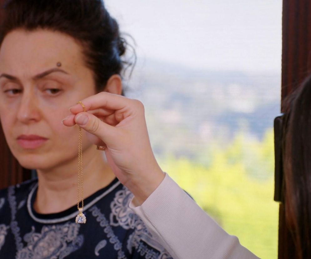 Cavidan z serialu Miłość i nadzieja patrzy na złoty naszyjnik z błyszczącym wisiorkiem, który podarowała jej Bahar w ramach przekupstwa po kłótni. Scena z tureckiego serialu Miłość i nadzieja na tle okna i zieleni. Więcej informacji o serialu znajdziesz na Super Seriale.