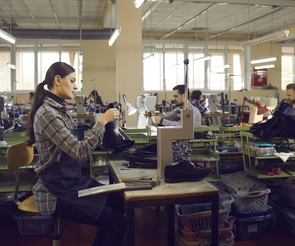 Pracownica w fartuchu w szwalni, w tle inni pracownicy i maszyny do szycia. Kobieta trzyma w rękach czarne obuwie, pracując w zakładzie produkcyjnym. Taki obraz może ilustrować temat pułapki w pracy, o której przeczytasz na Poradnik Zdrowie.