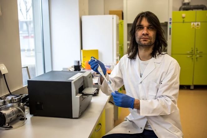 Dr hab. Piotr Rzymski z Uniwersytetu Medycznego w Poznaniu, ubrany w biały kitel i niebieskie rękawiczki, pracuje w laboratorium z pipetą automatyczną. Opisuje, czy wariant Mu koronawirusa jest powodem do obaw, o czym przeczytasz na Poradnik Zdrowie.