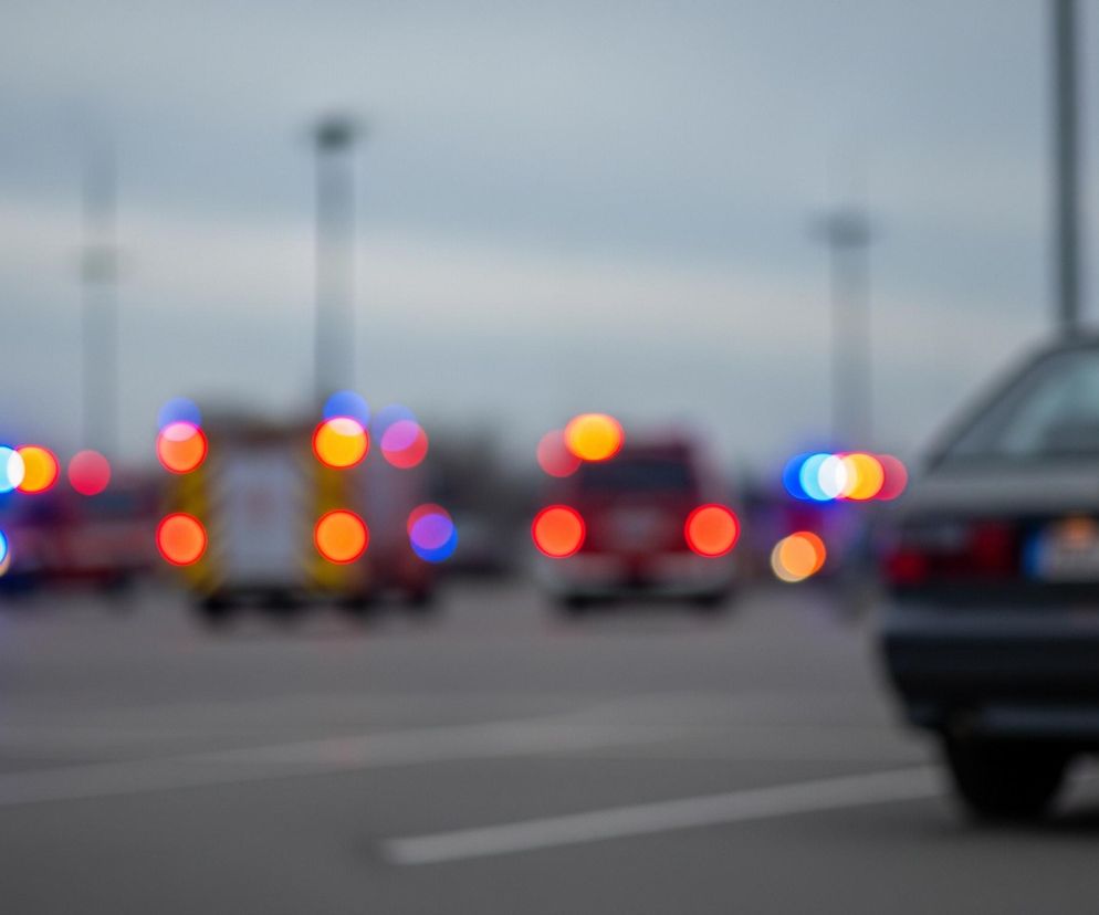 W prawym dolnym rogu widoczna jest tylna część ciemnego samochodu osobowego z wyraźnymi detalami, takimi jak tylne światła i tablica rejestracyjna. Większość obrazu jest nieostra, ukazując szereg jaskrawych, rozmytych świateł w różnych kolorach – głównie czerwonym, niebieskim i pomarańczowym – rozsianych po środku kadru, tworzących efekt bokeh. Światła te pochodzą od pojazdów służb ratunkowych, które są niewyraźne, ale można dostrzec ich kształty i kolorystykę, taką jak czerwony wóz strażacki i żółto-biały pojazd. Tło stanowi szare, zachmurzone niebo oraz niewyraźne sylwetki słupów oświetleniowych.