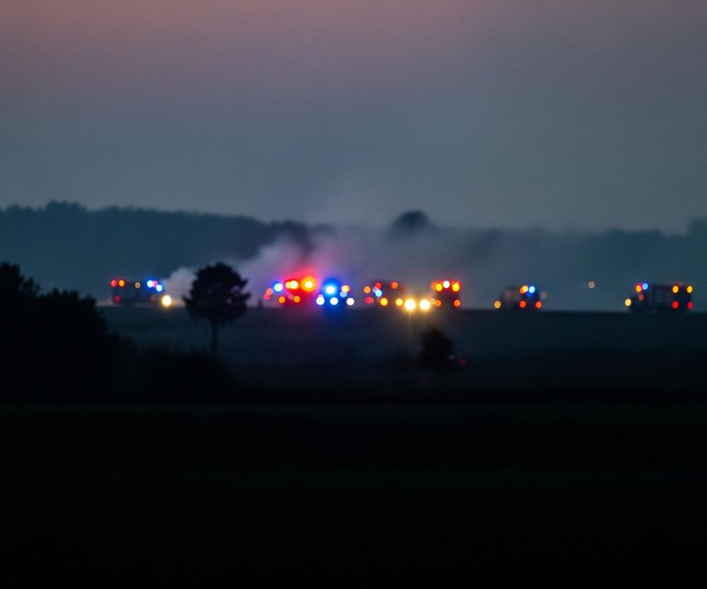 Na pierwszym planie, w dolnej części kadru, rozciąga się ciemne, niewyraźne pole, na którym po lewej stronie widać dwa rozmyte, ciemne kształty drzew. Dalej, na linii horyzontu, wyróżnia się szereg rozświetlonych punktów w kolorach czerwonym, niebieskim i pomarańczowym, które wydają się pochodzić z pojazdów ratunkowych lub ostrzegawczych. Za nimi unosi się biały dym lub mgła, rozświetlona światłami. Tło stanowi zamazana linia ciemnych drzew, a nad nią rozciąga się niebo o stopniowo zmieniających się barwach, od ciemnego granatu u dołu do delikatnego różu i fioletu u góry.