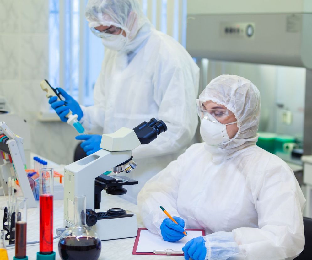 Dwoje naukowców w białych kombinezonach ochronnych, maskach i niebieskich rękawiczkach, pracuje w laboratorium. Osoba na pierwszym planie notuje przy mikroskopie, podczas gdy druga trzyma pipetę z niebieską substancją. Scena ilustruje badania nad koronawirusem, o których więcej przeczytasz na portalu Poradnik Zdrowie.