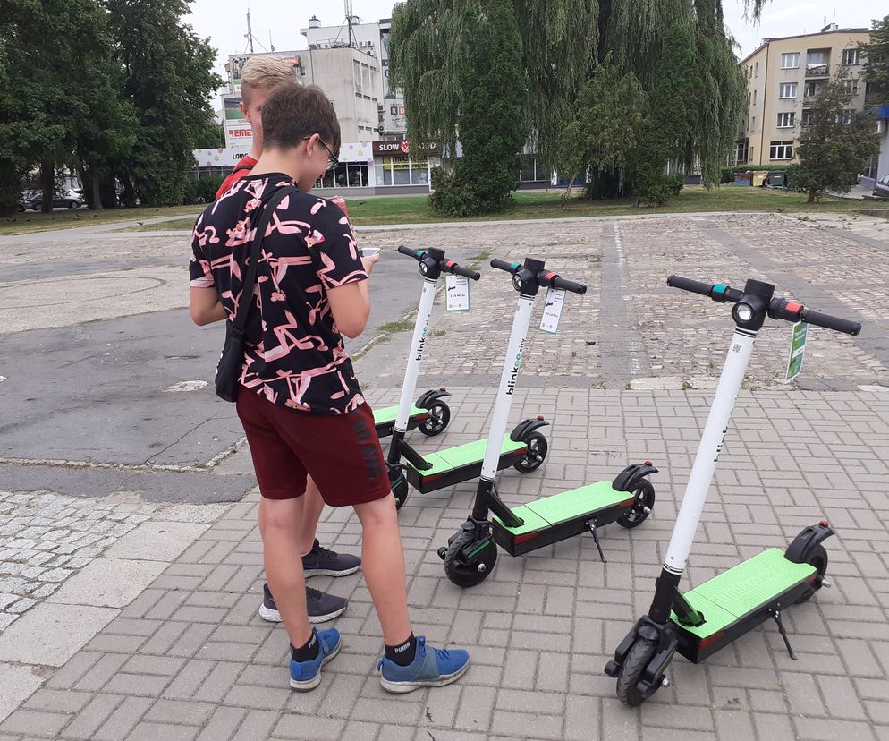 Dwie osoby stojące obok trzech wypożyczalnych hulajnóg elektrycznych na brukowanym placu. Młodzieńcy przeglądają telefony, prawdopodobnie przygotowując się do skorzystania z pojazdów. W tle budynki i drzewa. Sopot planuje strefy bez hulajnóg, o czym przeczytasz na Super Biznes.