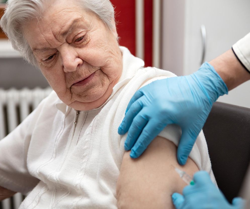 Starsza kobieta w jasnym kardiganie otrzymuje zastrzyk w ramię od osoby w niebieskich rękawiczkach, co symbolizuje darmowe szczepienia dla seniorów na RSV. Dowiedz się więcej o profilaktyce i oszczędnościach na Poradnik Zdrowie.