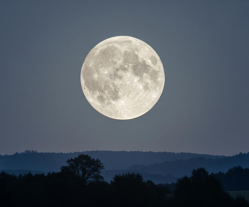 Na zdjęciu dominującym elementem jest jasny, prawie biały, pełny księżyc, umiejscowiony centralnie na niebie. Jego powierzchnia ukazuje wyraźne kratery i zróżnicowaną teksturę. Niebo wokół księżyca ma stonowany, ciemnoniebieski odcień. Poniżej, w dolnej części kadru, rozciąga się pasmo wzgórz i lasów, widoczne jako ciemne sylwetki w odcieniach czerni i głębokiego granatu, tworząc warstwową perspektywę.