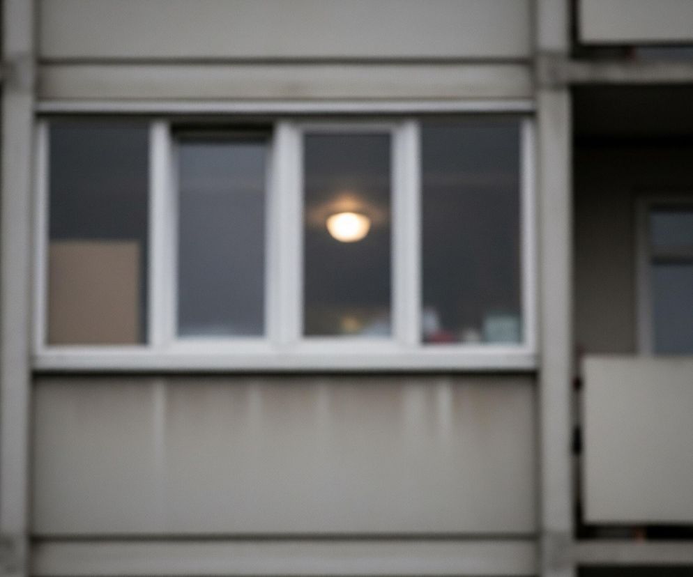 Środkowa część obrazu to duże, poziome okno z czterema białymi ramami, zza którego prześwituje rozmyte, żółto-białe światło żarówki. Po bokach okna widoczne są fragmenty dwóch balkonów o betonowych balustradach w szarym kolorze. Całość kompozycji, poza rozświetloną żarówką, charakteryzuje się rozmytą i nieostrą scenerią.