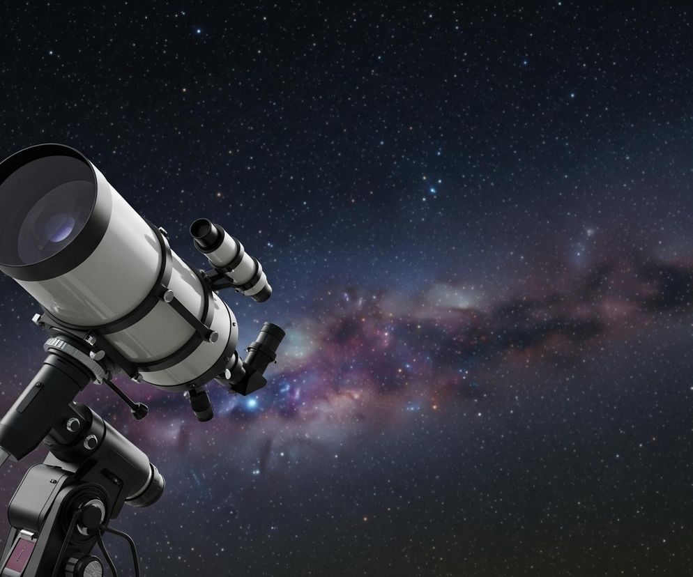 W lewej części obrazu znajduje się duży teleskop astronomiczy w kolorze białym i czarnym, skierowany ukośnie w górę, w prawą stronę. Tubus teleskopu jest biały, z czarnymi pierścieniami mocującymi i osłoną obiektywu; widoczny jest również mniejszy, czarny lunetka celownicza przymocowana do głównego tubusu. Teleskop zamontowany jest na solidnym, czarnym statywie i montażu, częściowo widocznym u dołu po lewej stronie. Tło stanowi ciemne niebo pełne gwiazd, z wyraźną, rozmazaną smugą Drogi Mlecznej w odcieniach fioletu, różu i błękitu, rozciągającą się od środka ku prawej górze. Gwiazdy są różnej wielkości i jasności, tworząc gęsty, kosmiczny pejzaż.