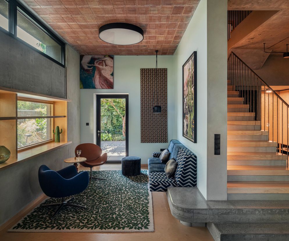 Wnętrze Domu Zosi, projektu IFAgroup w Gdańsku, z betonową ścianą, drewnianymi schodami i ceglanym sufitem. Na pierwszym planie niebieski fotel, sofa w zygzaki i zielony dywan, ukazujące ciekawe rozwiązania wnętrzarskie, o których przeczytasz na Architektura Murator Plus.