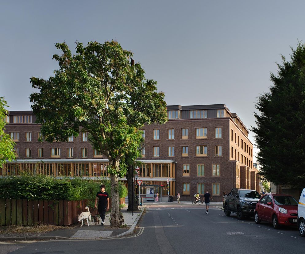 Zewnętrzny widok na Appleby Blue Almshouse w Londynie, nowoczesny budynek z cegły z drewnianymi detalami, otoczony zielenią i drzewami. Na ulicy widać przechodniów z psami i zaparkowane samochody. Więcej o architekturze tego typu na Architektura Murator Plus.