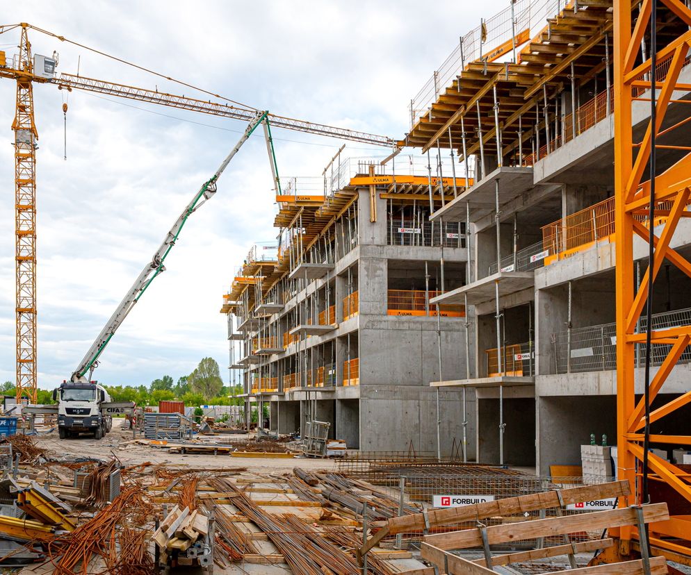 Wielki plac budowy z budynkami w szkieletowej konstrukcji, betoniarką z pompą do betonu oraz wysokimi żurawiami, symbolizujący rewolucję w prawie budowlanym i problemy w urzędach. O zmianach i ich wpływie na rynek nieruchomości przeczytasz na Super Biznes.