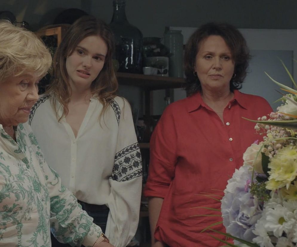 Teresa Lipowska jako Barbara, Dominika Kachlik jako Franka i nieznana kobieta w czerwonej koszuli, patrzące na bukiet kwiatów, w domu Mostowiaków. Więcej o ich losach dowiesz się na portalu Super Seriale.