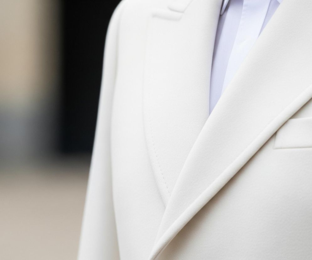 Zbliżenie przedstawia prawą część górnej partii ciała osoby ubranej w jasny garnitur. Dominują odcienie bieli i kremu widoczne na marynarce z wyraźnie zaznaczonym kołnierzem i klapą. Pod marynarką widać jasną koszulę. Lewa strona zdjęcia jest rozmyta, ukazując niewyraźne, ciemniejsze tło.