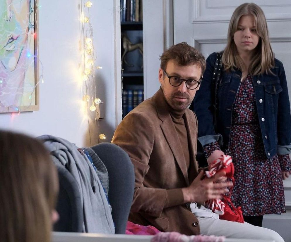 Darek Hoffman (Mateusz Rusin) siedzi na kanapie w domu, trzymając w rękach czerwony strój kąpielowy, podczas gdy Lenka (Olga Cybińska) stoi za nim, spoglądając zaniepokojona. Ta scena ukazuje napiętą atmosferę w M jak miłość, o której możesz przeczytać więcej na Super Seriale.