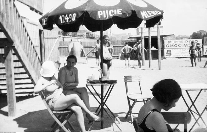 Plażowicze podczas wypoczynku pod parasolem z napisem "Pijcie mleko". W tle widoczna rekalma z napisem "Pikcie piwo Okocimskie". Zdjęcie zrobione w 1931 roku 