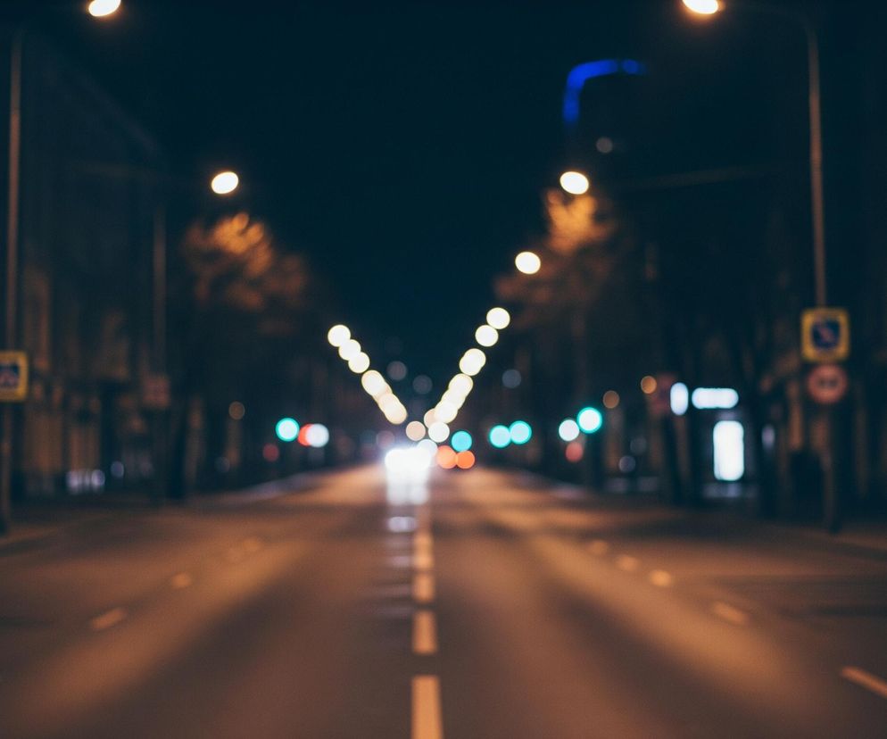 Droga asfaltowa, z białymi przerywanymi liniami oddzielającymi pasy ruchu, rozciąga się w głąb obrazu, rozświetlona przez światła uliczne i reflektory samochodów. Po obu stronach drogi widoczne są niewyraźne sylwetki budynków oraz drzew, a także liczne, rozmyte punkty świetlne o barwie żółtej, czerwonej i turkusowej, tworzące efekt bokeh. Żółte znaki drogowe w kształcie kwadratów z czarnymi symbolami, a także okrągłe znaki z czerwonym obramowaniem, są rozłożone wzdłuż jezdni po obu stronach. Horyzont dominuje ciemne, nocne niebo, z delikatnym niebieskim poświatą w prawym górnym rogu.